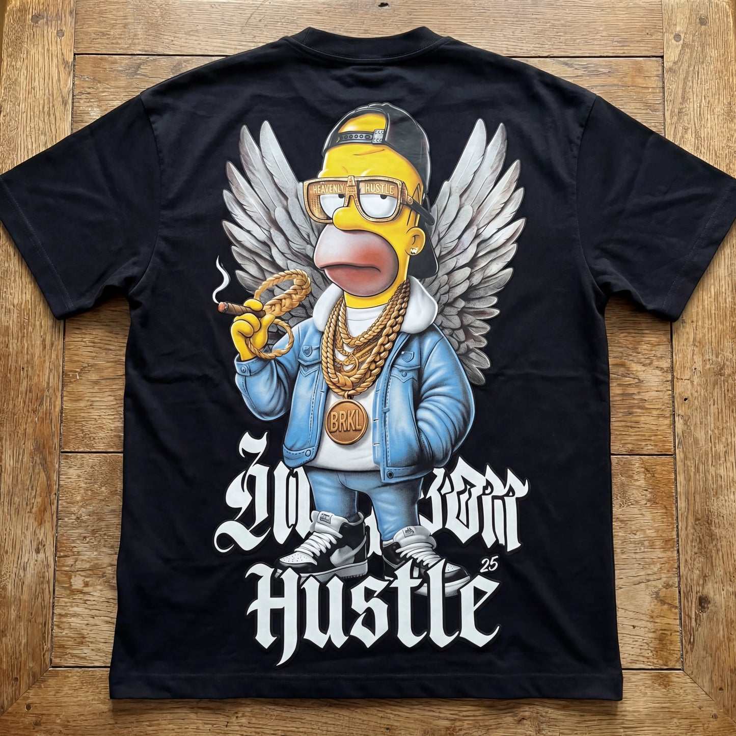 Black t-shirt with a graphic of a character wearing a blue jacket, gold chains, and sneakers on a wooden surface.